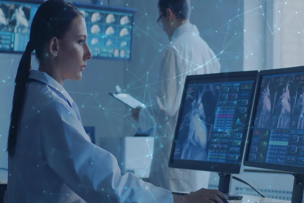 Woman in a lab coat works on two computer screens, focused on tasks related to Healthcare IT Services