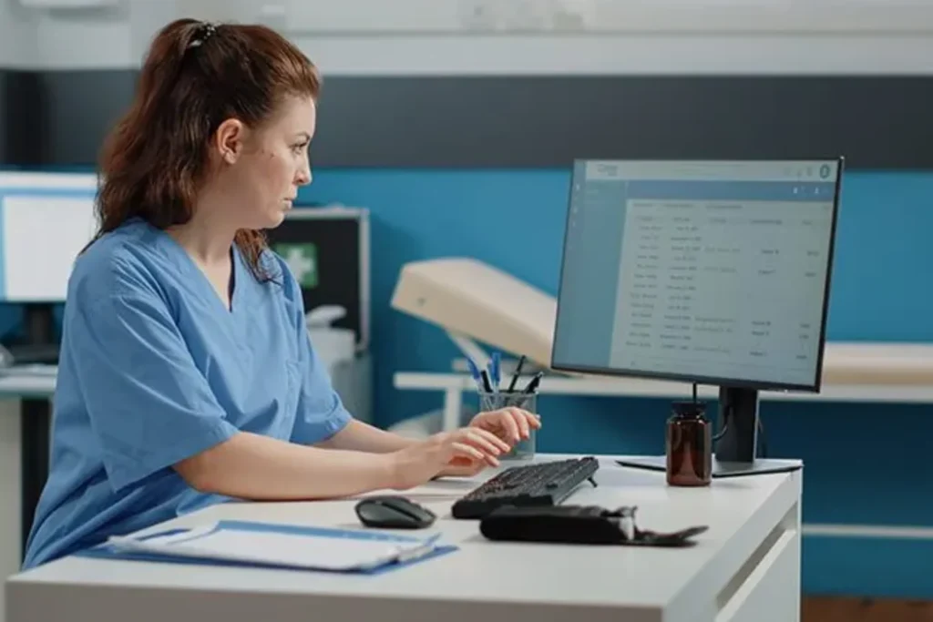 Woman in scrubs sits at a desk with a computer, focused on managing revenue cycle systems for healthcare practices
